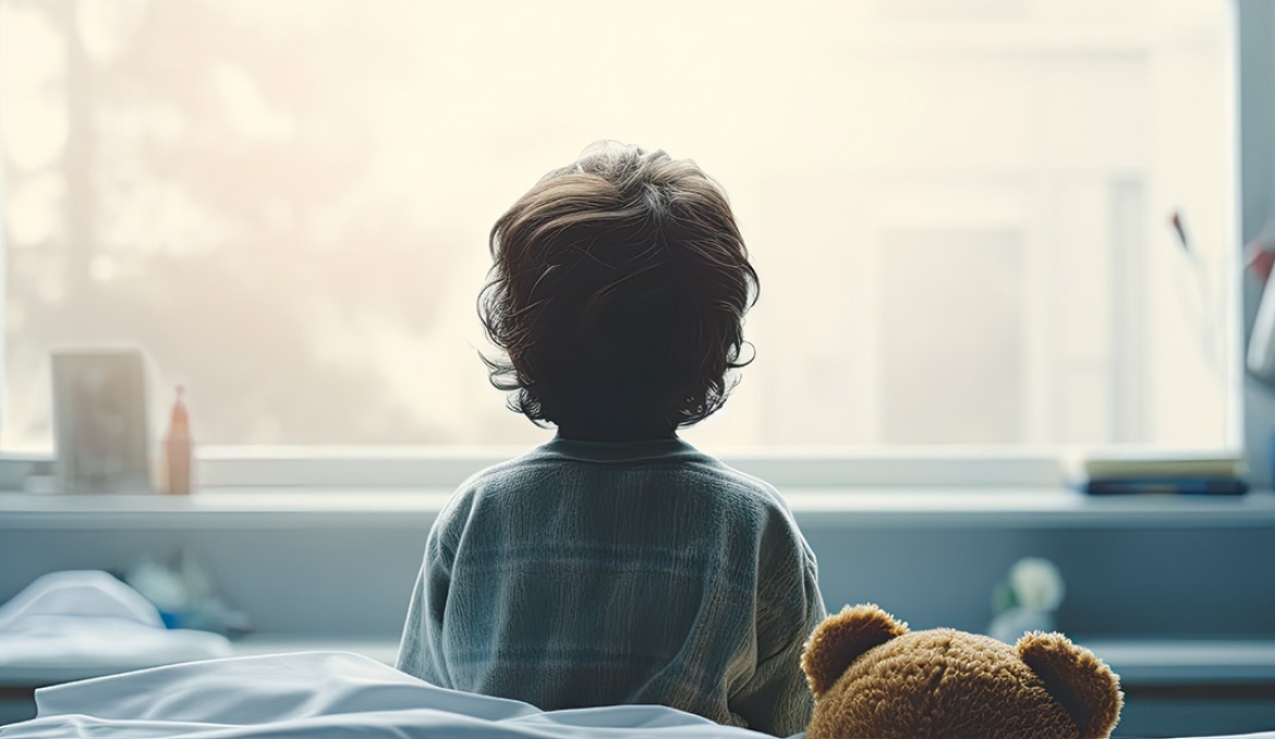 Ein Kind sitzt mit einem Teddy auf einem Bett und schaut aus dem Fenster. 